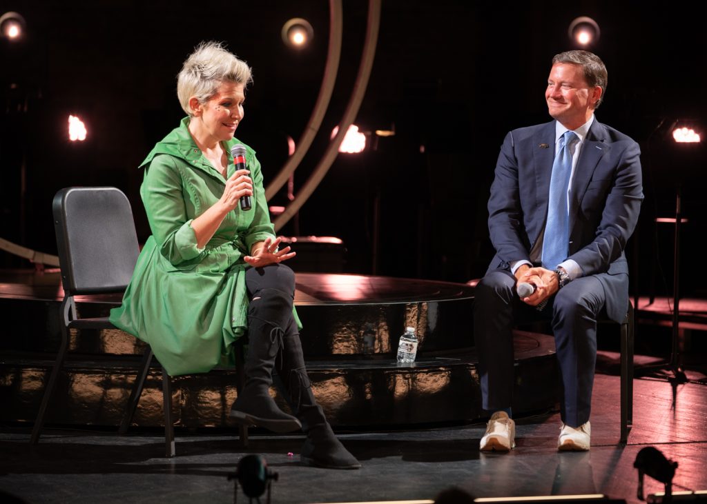 Joyce speaking on stage while someone sits and listens.