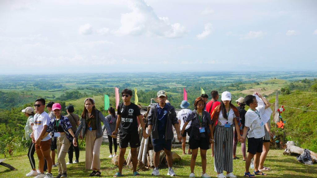 Students holding hands in a circle on top of a hill. They are facing towards the outside view.