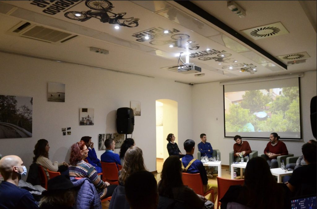 People presenting with screen behind them, audience seated in front.